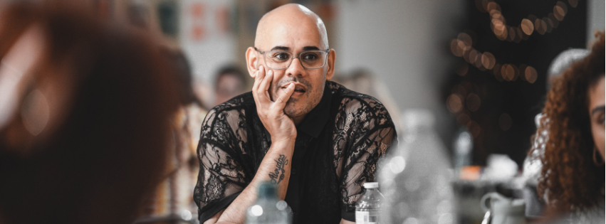 A non-binary person with a shaved head and glasses looks pensively across a table. The image uses shallow focus to isolate them from the busy background of a community event. They are wearing a black lace shirt and have tattoos visible on their forearms.