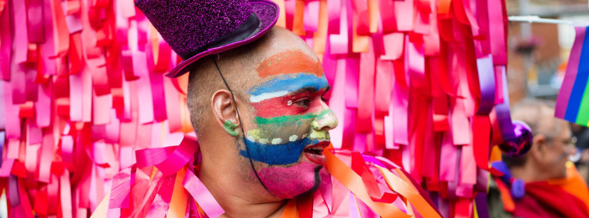 A close up of a man at a Pride parade, with a brightly-painted face and wearing a purple glittery top hat, against a background of pink