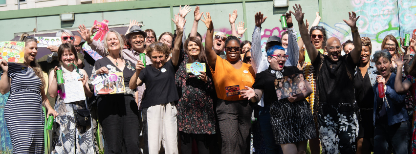The image shows a large group of people standing facing the camera in an urban garden in the sunshine. They are smiling and raising their hands in the air.
