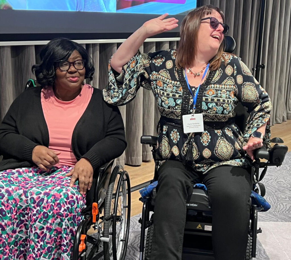 Two women sit in wheelchairs in front of a projection screen. The woman on the left is Black and is smiling, wearing a funky coloured leopard print skirt and peach top. The woman on the right is white and wears a boho style blouse and black trousers. She is smiling and reaching her hand above her shoulder.