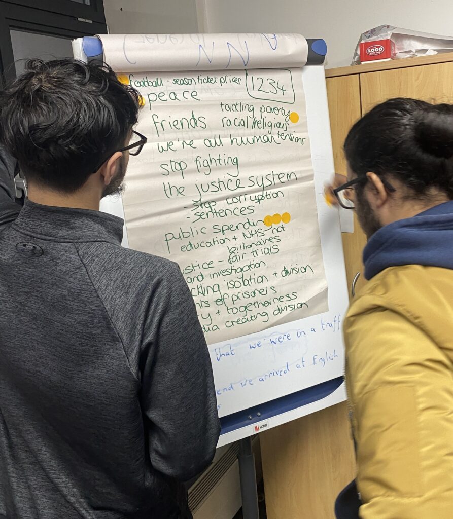 A photograph of two teenage boys standing in front of a flipchart. They are adding yellow dot stickers to the flipchart, on which is a handwritten list of different topics relating to society.