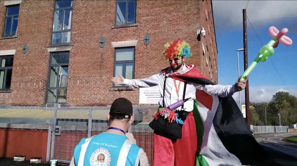 A young person in a blue Army of Kindness vest has their back to the camera, as they interview a clown on stilts in a rainbow wig and holding a balloon model of a flower.