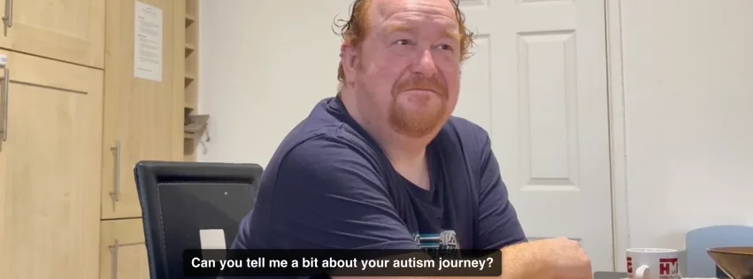 Image is a screen capture from a video interview. The subject is a white man with ginger hair and a goatee beard, wearing a navy tshirt and looking relaxed. A closed caption on the bottom of the image reads "Can you tell me a bit about your autism journey?"