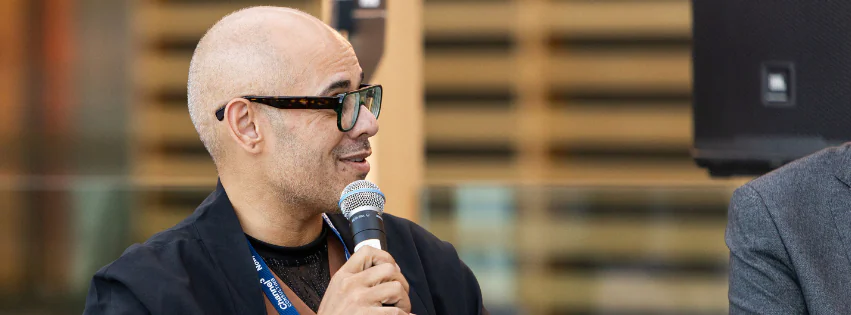 A person with a shaved head and glasses talks into a microhpone