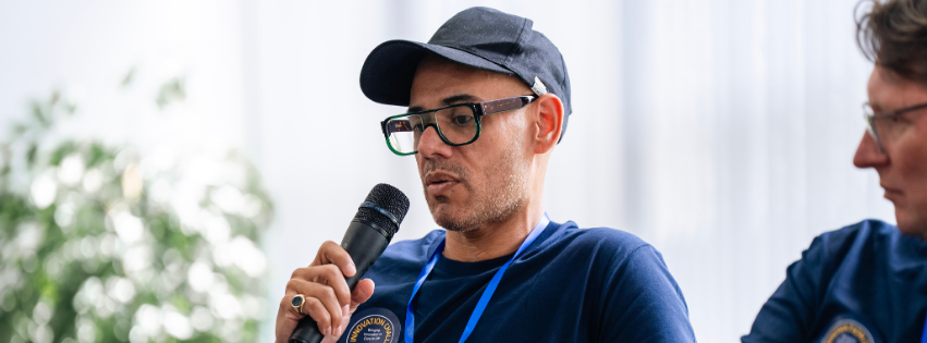 A non binary person in thick rimmed glasses and a baseball hat holds a microphone and looks serious.